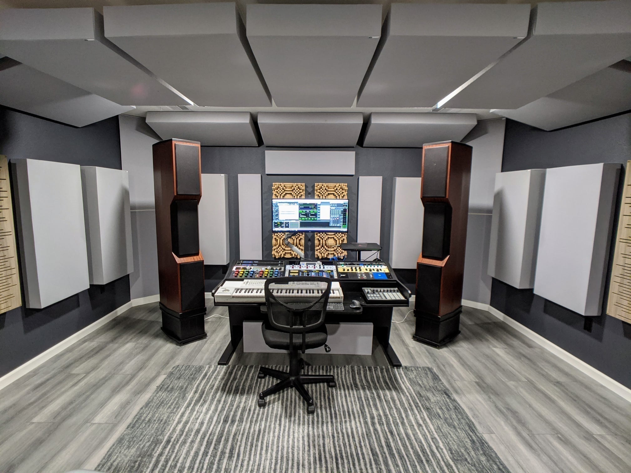 A fully treated mix position with GIK Acoustics acoustic panels and ceiling clouds, large floor-standing speakers positioned in an equilateral triangle with the mix desk, and Gotham Primitive Root Diffusers visible on the rear wall.