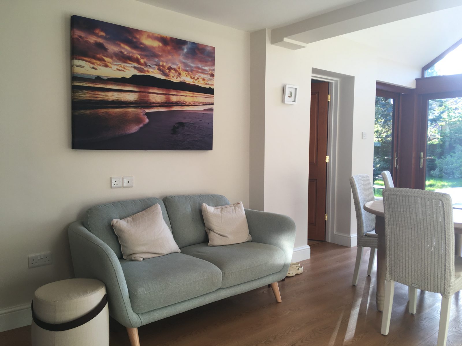 GIK Acoustics Acoustic Art Panel featuring a sunset beach landscape photograph, installed on the wall of a residential living room above a sofa.