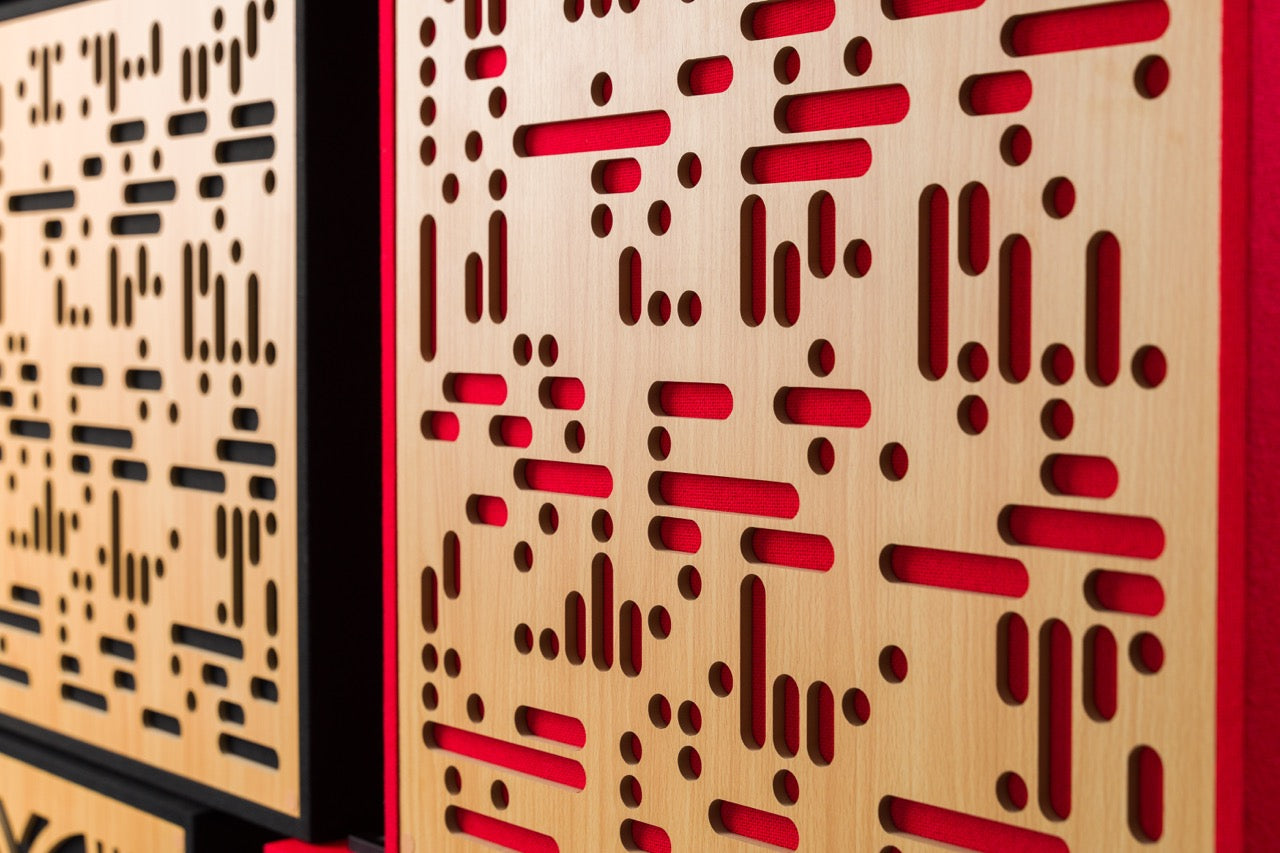 Close-up of GIK Acoustics SoundBlocks wood diffusion panel with a geometric cut pattern revealing red fabric beneath, paired with a black-framed panel in the background